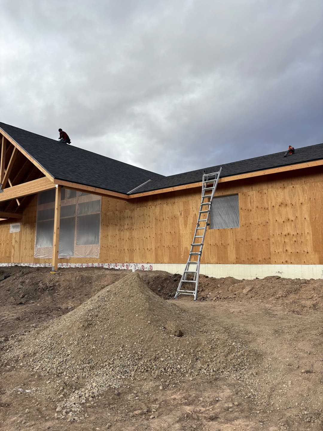 Workers installing a roof on a wooden house under cloudy skies, with a ladder and gravel pile nearby.