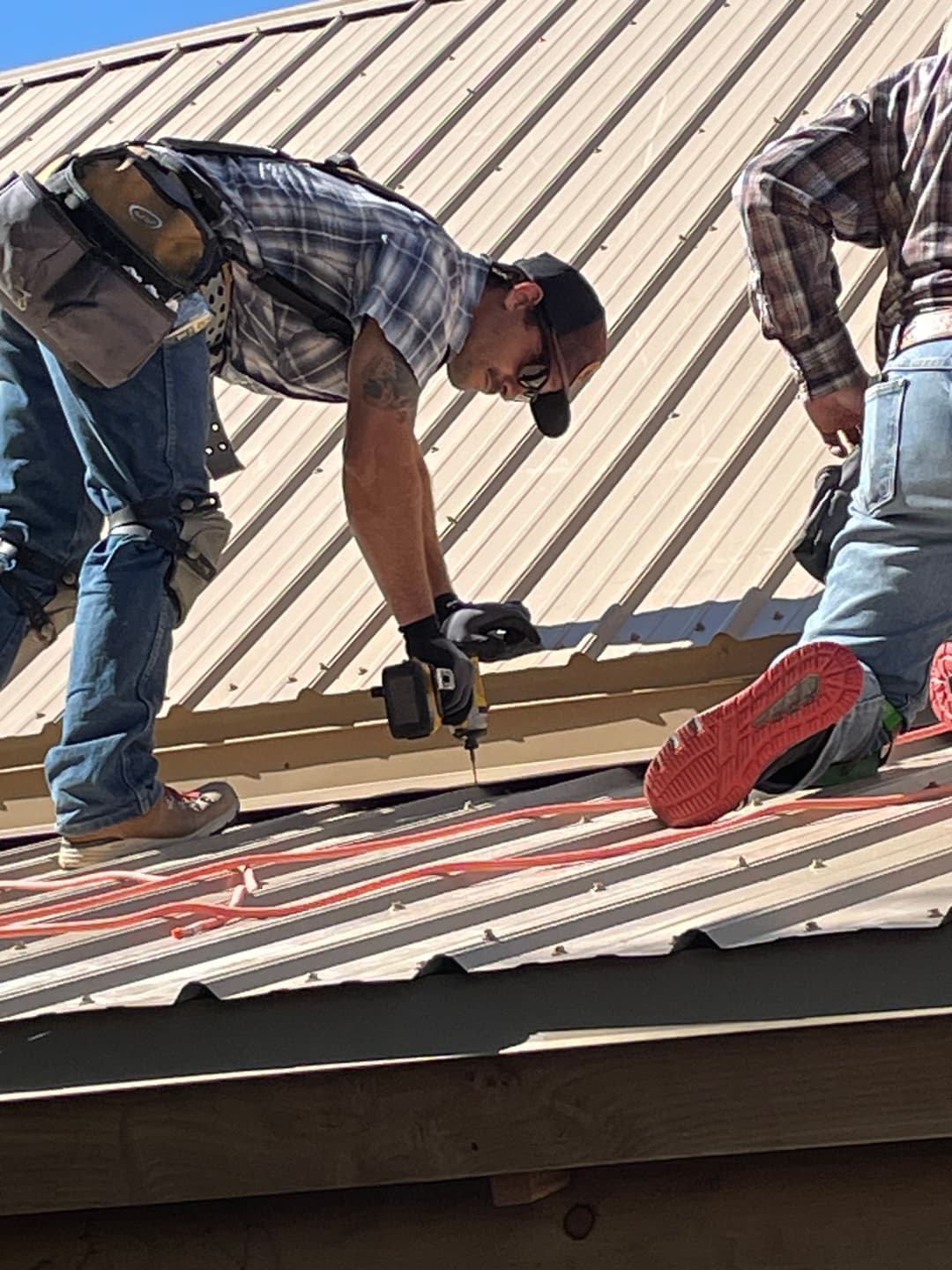 Roofer installing panels on a metal roof with tools and safety gear on a sunny day.